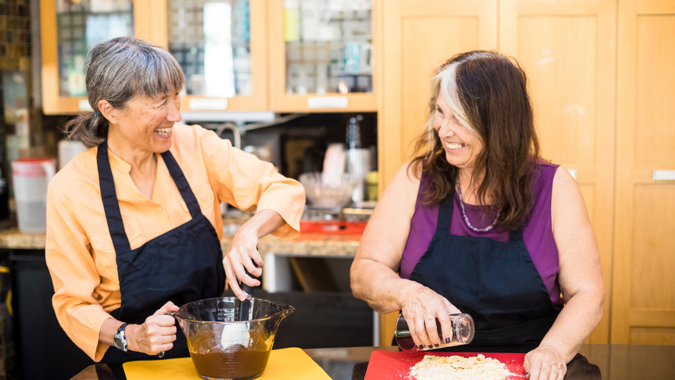 two senior women smile at each other while they cook separately two senior women smile at each other while they cook separately